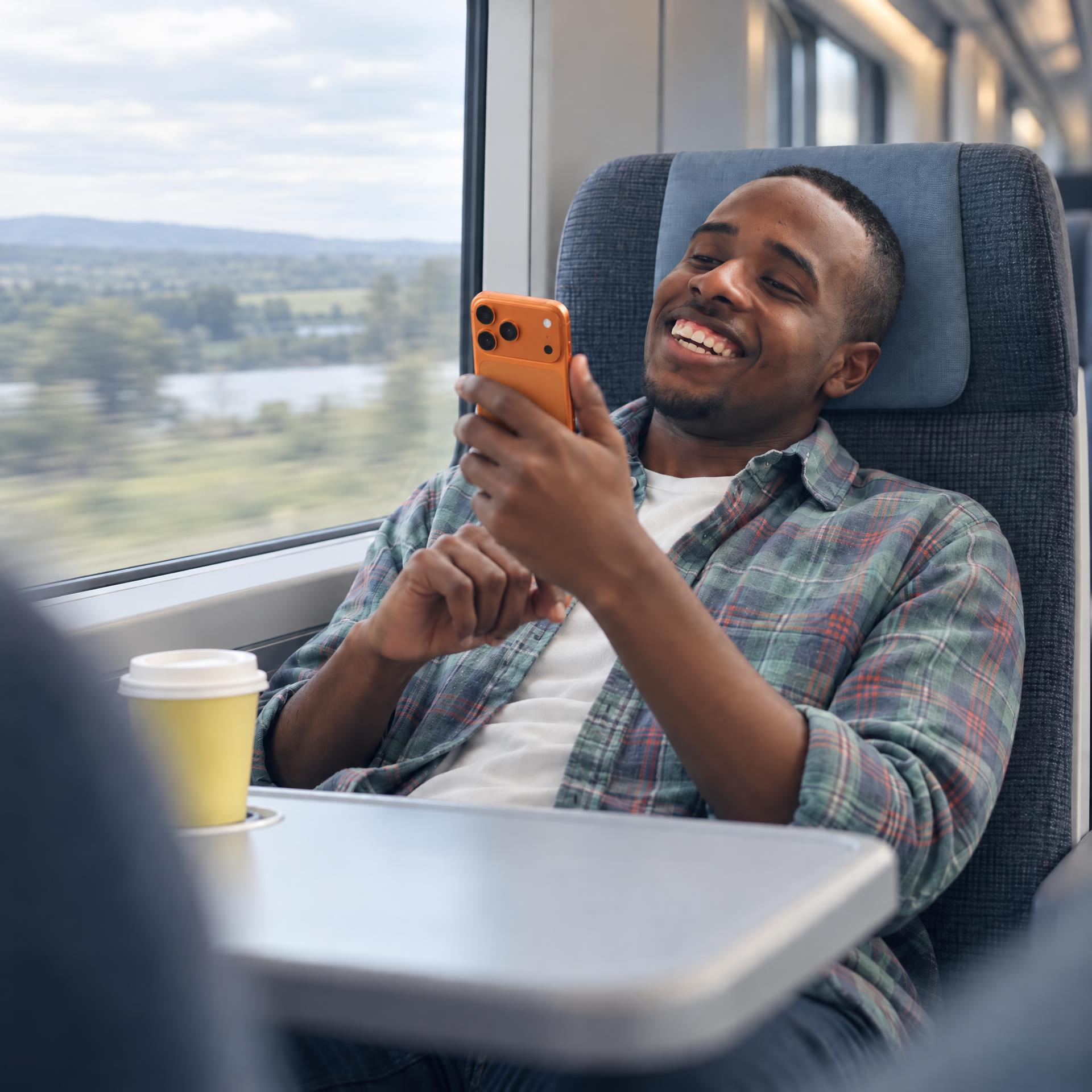 Ein junger Mann sitzt entspannt in einem Zugabteil und hält ein orangefarbenes Smartphone in der Hand. Auf dem Tisch vor ihm steht eine gelbe Kaffeetasse, während er aus dem Fenster auf die vorbeigleitende Landschaft blickt.