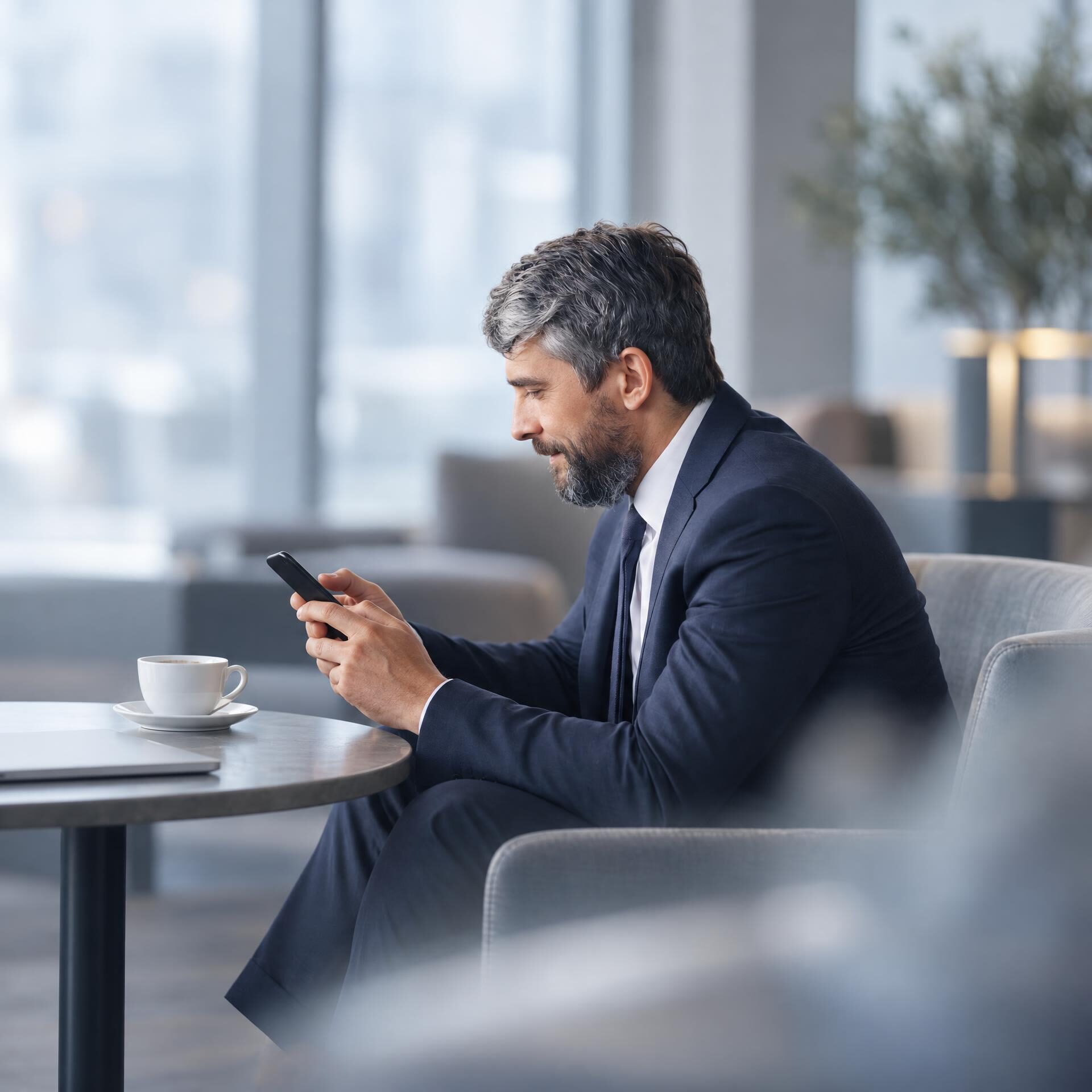 Ein Mann in einem Anzug sitzt in einem modernen Raum und schaut auf sein Smartphone. Auf dem Tisch vor ihm steht eine Tasse und eine Untertasse.